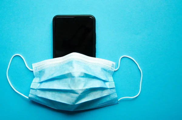 Siyah bir akıllı telefondaki mavi tıbbi maske. Koruma, akıllı telefonun dezenfeksiyonu. Maske, mavi, siyah, pembe, sarı arka plandaki akıllı telefondan giyinmiş. Uzayı kopyala COVID-19