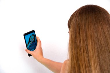 Ekose gömlekli, uzun saçlı genç bir kız beyaz arka planda duruyor ve elinde bir akıllı telefon tutuyor. Kız selfie çekiyor. Kızın ön ve arka vizeleri. Dudaklar eğik