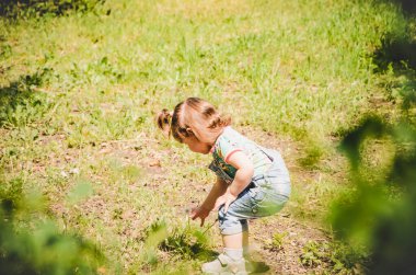 Mayıs 2020. Kiev Ukrayna. Parkta oynayan küçük bir kız, güneşli bir günde kot tulumuyla çiçek topluyor ve üzerinde de parmak izi olan bir tişört giyiyor.