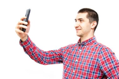 Beyaz arka planda akıllı telefonu olan ekose gömlekli genç bir adam. Adam akıllı telefona bakar ve selfie çekerken telefonu başının üstünde tutar.