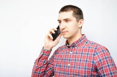 Beyaz arka planda akıllı telefonu olan ekose gömlekli genç bir adam. Adam akıllı telefonu kulağından tutup, telefonla konuşuyordu. Bir telefon konuşmasında öfke, öfke, şaşkınlık, dikkat.