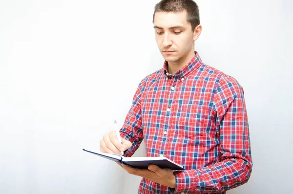 Beyaz arka planda akıllı telefonu olan ekose gömlekli genç bir adam. Adam elinde bir defter tutuyor ve bilgi, görev ve hatırlatmalar yazıyor.