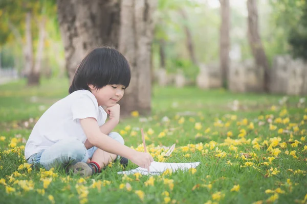 Liten asiatisk pojke sitter under trädet och ritning i anteckningsboken — Stockfoto