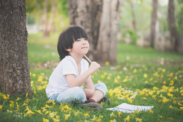 Liten asiatisk pojke sitter under trädet och ritning i anteckningsboken — Stockfoto