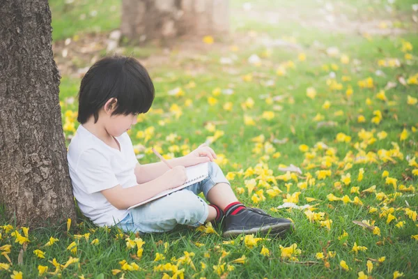 Liten asiatisk pojke sitter under trädet och ritning i anteckningsboken — Stockfoto