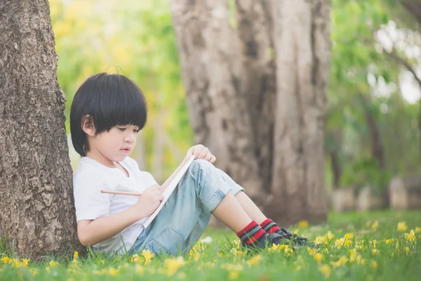 Liten asiatisk pojke sitter under trädet och ritning i anteckningsboken — Stockfoto