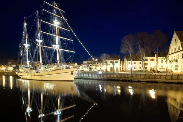 セーリングボートは、リトアニアのクライペダの古い町に停泊または係留され、夜のライトが点灯します。中世の町や穏やかな海でのBarquentine反射. — ストック写真