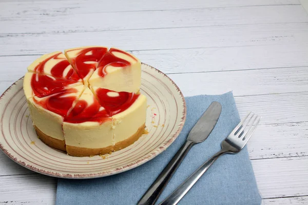 Tabakta cheesecake, mavi masa örtüsünde çatal ve bıçak beyaz ahşap arka planda izole edilmiş.