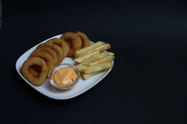 Soğan halkaları, patates kızartması ve siyah arka planda izole edilmiş beyaz tabakta bir kase peynir sosu. Resim kopyalama alanı içeriyor