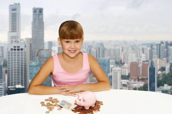 一个可爱的小女孩 坐在城市背景的桌子上 手里拿着硬币和小猪银行 为教育基金存钱 商业概念 — 图库照片