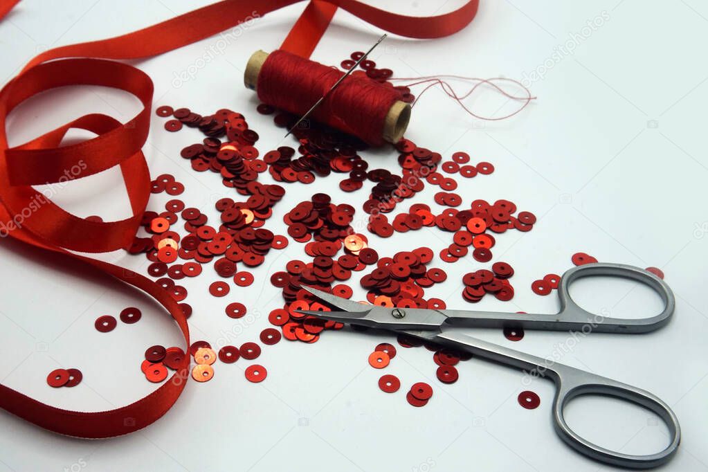 Red sequins, spool of red thread, needle, scissors and red ribbon on white background.