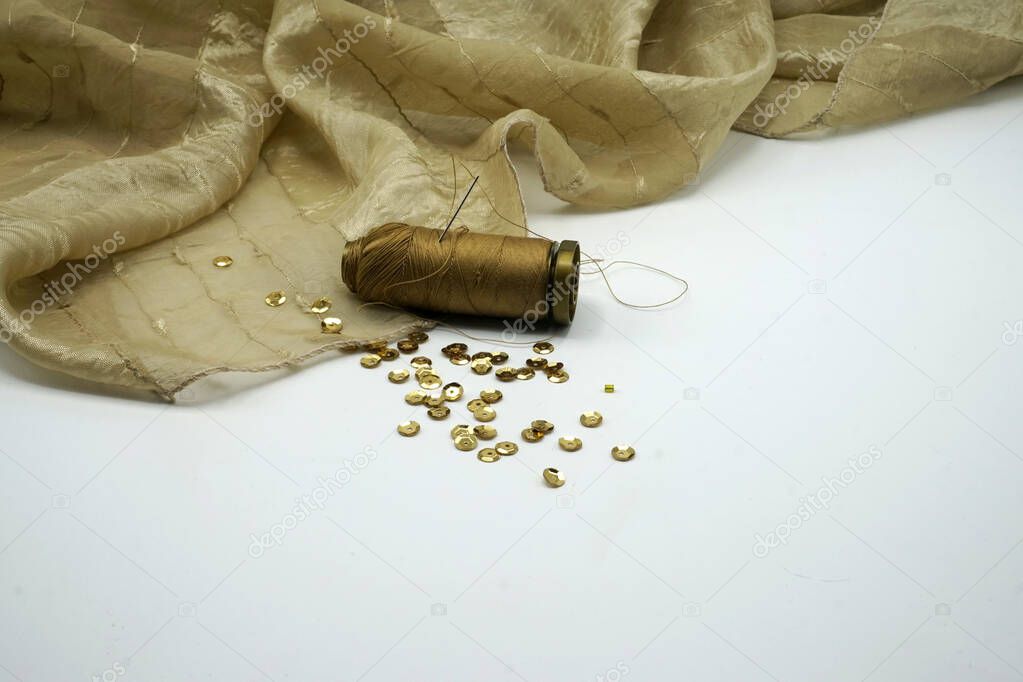 Gold sequins, spool of golden thread, needle on rippled gold silk background. Sewing and decorating accessories used for embellishment of dresses.