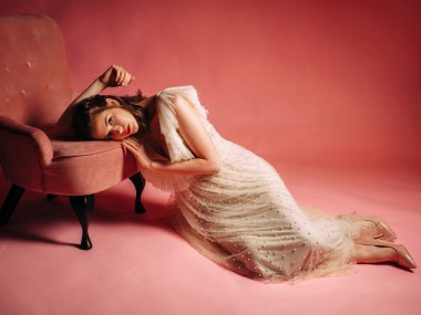 Somon arka planında fildişinden yapılmış pohpohlayıcı elbisesi olan güzel esmer kadın. Stüdyoda pembe koltuklu harika bir gelin. Moda çekimi..