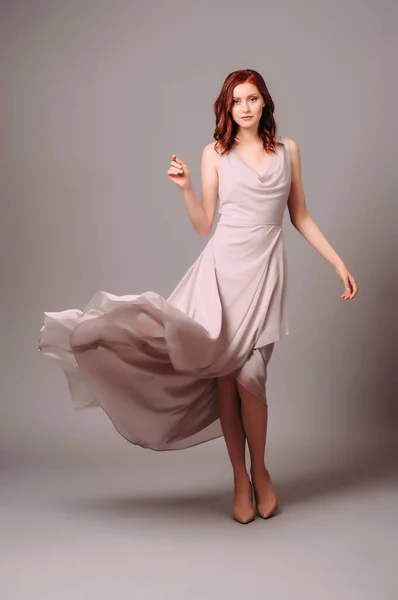 Toz pembe moda görünüş. Yazın kızıl saçlı genç bir kadının güzel portresi çırpınan elbisesi ve gri arka planda yüksek topuklu ayakkabıları stüdyoda.. 