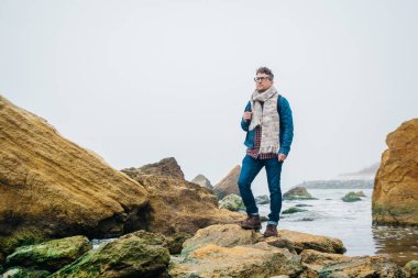 Dalgalar ile güzel bir denize karşı bir kaya üzerinde duran bir sırt çantası ile Adam gezgin, dünya çapında harika bir yolculuk sırasında sakin bir okyanus yakınında poz şık bir hipster çocuk.