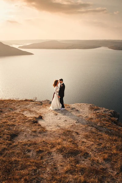 Increíble pareja de boda, novia y novio tomados de la mano en un fondo de montañas y ríos. Linda chica en vestido blanco, los hombres en traje de negocios. Montañas paisaje y ríos en la puesta del sol. Concepto de —  Fotos de Stock