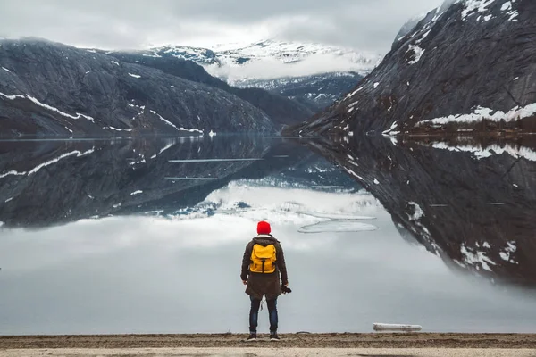 Ταξιδιώτης με ένα κίτρινο σακίδιο φορώντας ένα κόκκινο καπέλο που στέκεται στο φόντο των βουνών και της λίμνης απολαμβάνοντας το τοπίο. Σχέδιο ταξιδιωτικού τρόπου ζωής. Ρίξε από πίσω. — Φωτογραφία Αρχείου