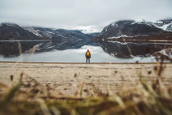 Ταξιδιώτης με ένα κίτρινο σακίδιο φορώντας ένα κόκκινο καπέλο στέκεται στο φόντο των βουνών και της λίμνης απολαμβάνοντας το τοπίο στο Lofoten, Νορβηγία. Σχέδιο ταξιδιωτικού τρόπου ζωής. Ρίξε από πίσω. — Φωτογραφία Αρχείου