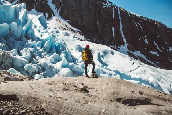 Reisender mit gelbem Rucksack und rotem Hut, der auf einem Felsen auf dem Hintergrund eines Gletschers, der Berge und des Schnees steht. Travel Lifestyle Konzept. Schuss von hinten — Stockfoto