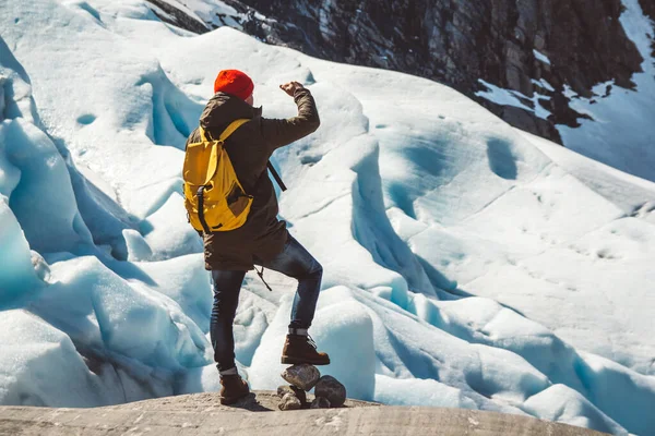 Hombre viajero con una mochila amarilla con un sombrero rojo de pie sobre una roca en el fondo de un glaciar, montañas y nieve. Concepto de estilo de vida. Dispara por la espalda — Foto de Stock