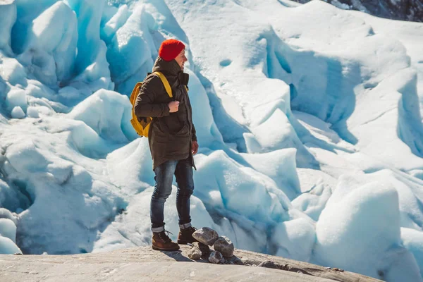 Reiziger met een gele rugzak met een rode muts op een rots op de achtergrond van een gletsjer, bergen en sneeuw. Reizen lifestyle concept — Stockfoto