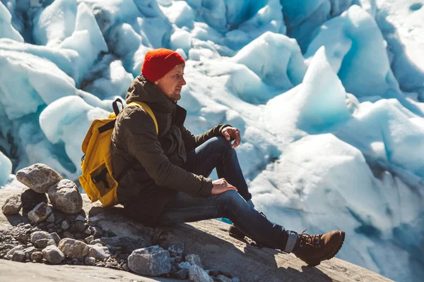 Hombre viajero con una mochila amarilla con un sombrero rojo sentado en una roca en el fondo de un glaciar y nieve. Concepto de estilo de vida — Foto de Stock