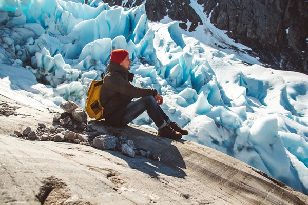 Hombre viajero con una mochila amarilla con un sombrero rojo sentado en una roca en el fondo de un glaciar y nieve. Concepto de estilo de vida — Foto de Stock