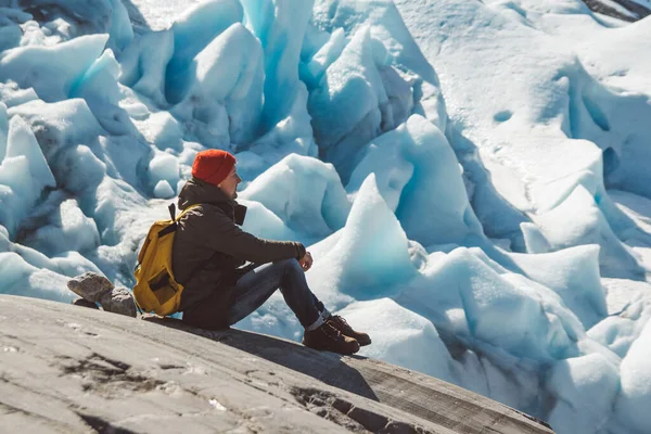 Homem viajante com uma mochila amarela usando um chapéu vermelho sentado em uma rocha no fundo de uma geleira e neve. Conceito de estilo de vida — Fotografia de Stock