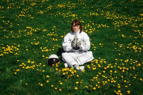 Beautiful girl astronaut without a helmet sits on a green lawn among flowers in a meditative position.