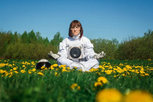 Schöne Astronautin ohne Helm sitzt auf einem grünen Rasen zwischen Blumen in einer meditativen Position. — Stockfoto