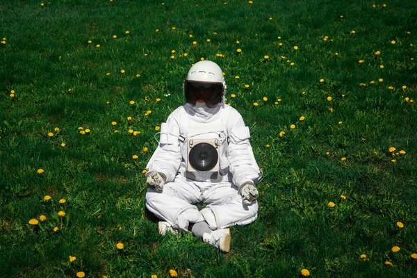 Futuristic astronaut in a helmet sits on a green lawn among flowers. Greetings to the camera