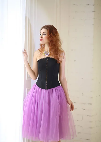 Beautiful red-haired girl in a purple tulle skirt and black corset is standing near the floor window — Stock Photo, Image