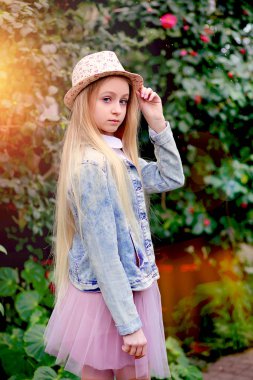 Uzun beyaz saçlı, şapkalı ve kot ceketli genç bir kız bahçede yürüyor. 