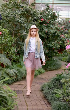 Uzun beyaz saçlı, şapkalı ve kot ceketli genç bir kız bahçede yürüyor.