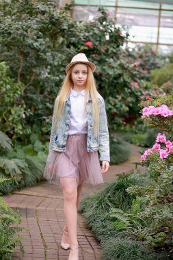 Uzun beyaz saçlı, şapkalı ve kot ceketli genç bir kız bahçede yürüyor. 