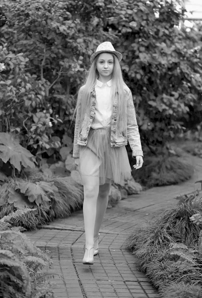 Молодая девушка с длинными белыми волосами в шляпе и джинсовой куртке гуляет по саду — стоковое фото