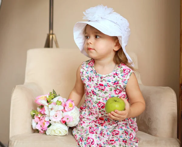 白い帽子をかぶった椅子に座っている女の子とリンゴの花のプリントのドレス — ストック写真