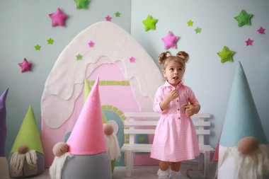 Pembe kareli elbiseli hoş beyaz kız parlak odasında büyük oyuncak cüceler arasında eğleniyor.