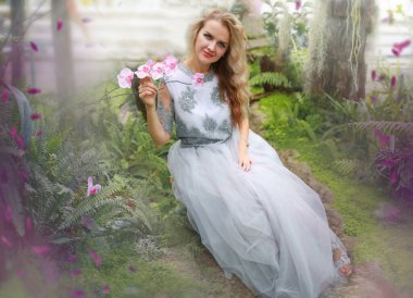 Yeşilliklerin arasında cam bir serada genç, beyaz, sarışın bir kız. Romantik gri elbiseli bir kızın sanatsal portresi.