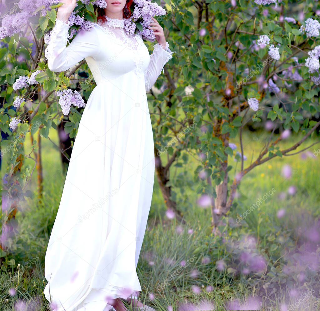 young beautiful red-haired Caucasian girl in a white dress enjoys lilac blossoms in spring park