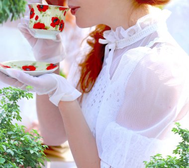 Beyaz elbiseli ve eldivenli güzel kızıl saçlı beyaz bir kız elinde fincan ve çay tabağı, bahçede bir piknikte çay içiyor, klasik tarzda.