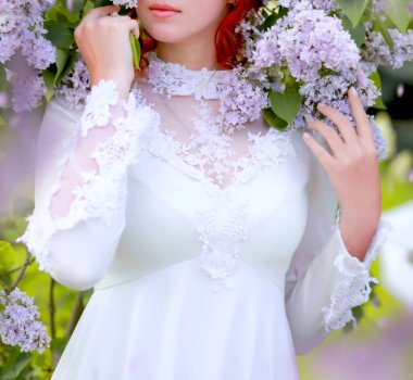 Beyaz elbiseli güzel kızıl saçlı beyaz kız bahar parkında leylak çiçeklerinin tadını çıkarıyor.