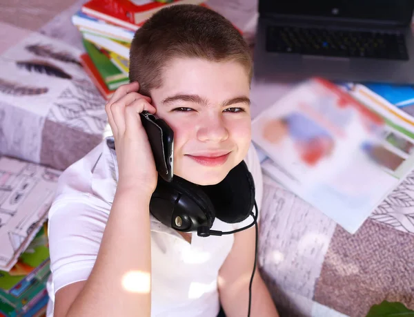 A young Caucasian boy in headphones is studying with a books and a laptop. Calls a friend on the mobile phone. Distance learning during a pandemic 2019-ncov. Insulation