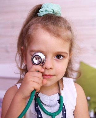 Küçük çekici beyaz kız doktoru oynuyor, bir gözünü steteskopla kapatıp gülümsüyor. Çocuklar işte. Sağlık kavramı. Salgın. Karantinaya alın. Yalıtım. Store coronavirus 2019-ncov