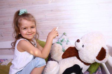 Kafkasyalı, maske takmış, elinde burun spreyi olan bir ayıyla doktorculuk oynayan küçük bir kız. Koronavirüs 2019-ncov 'a karşı karantina koruması. Ev yalıtımı