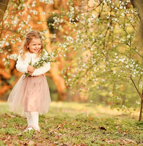一个漂亮可爱的小女孩在春天的植物园里 穿着一条飘扬的裸体裙子和一件白色的毛衫 头戴一束盛开的樱花 笑着玩乐 复活节和春季卡片 — 图库照片