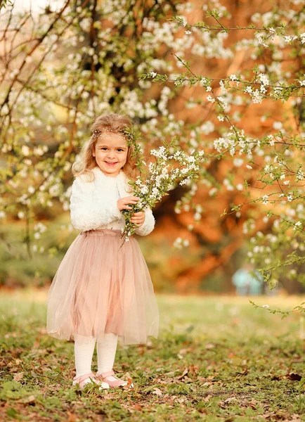 一个漂亮可爱的小女孩在春天的植物园里 穿着一条飘扬的裸体裙子和一件白色的毛衫 头戴一束盛开的樱花 笑着玩乐 复活节和春季卡片 — 图库照片