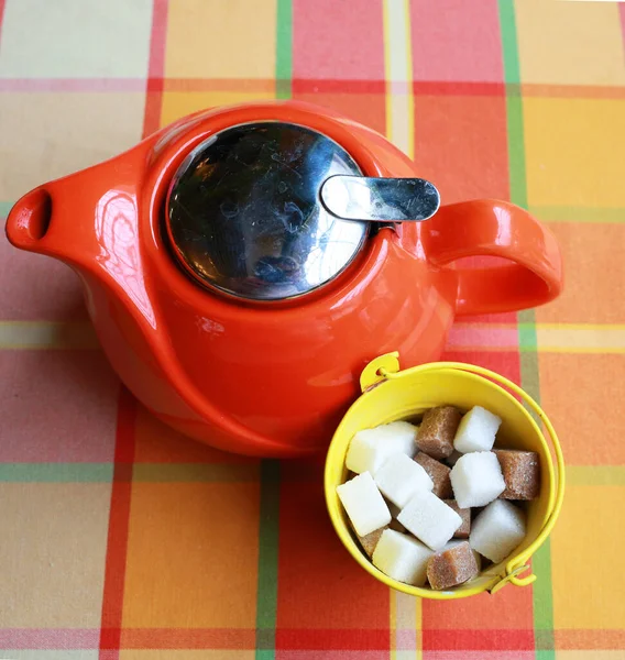 Red ceramic teapot and cubes white sugar and cane sugar isolated on a colored checkered tablecloth, top view. Tea ceremony. Healthy eating concept. Close-up
