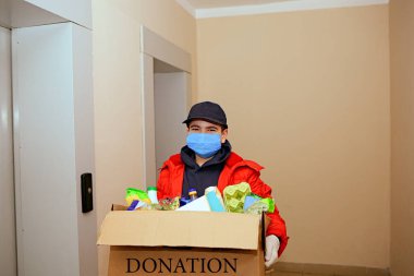 Koruyucu tıbbi maske takan gönüllü, genç, beyaz bir adam ve yiyecek dolu bir bağış kutusuyla eldiven. Yakın plan. Ev yalıtımı. Coronavirus 2019-nkov. Bağış. Teslimat. Yiyecekler. Gönüllü