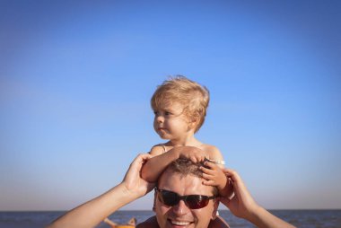 Babamın boynunda oturan mayolu mutlu küçük kız. Mavi gökyüzü ve denizin arka planı, tatil ve seyahat. Babalar Günü. Kızların günü. Aile günü. Küçük çocukların yanında dinlen.